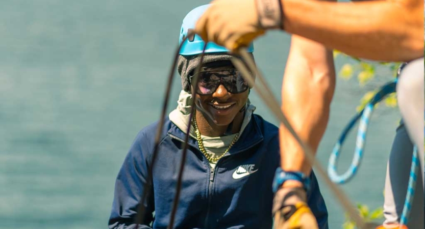 In the foreground, two hands handle a rope. Behind, a person wearing a helmet is smiling. They appear to be at high elevation. There is water behind them.